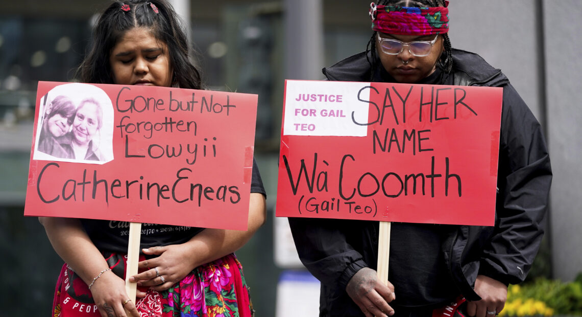 Two Native American protestors hold red signs with the names of Indigenous victims.