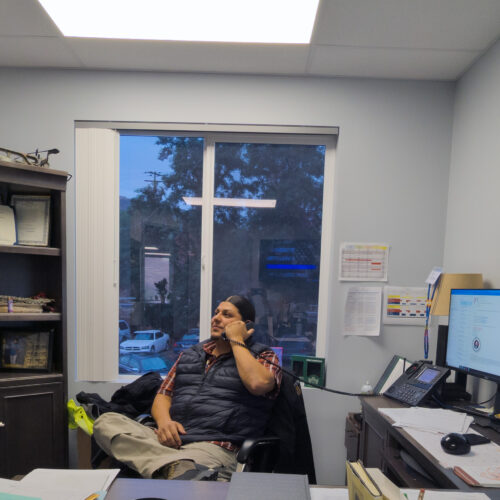 Leotis McCormack sits at his desk with double computer monitors and a brown shelf behind him, filled with knick-knacks. He holds a phone in his hand.