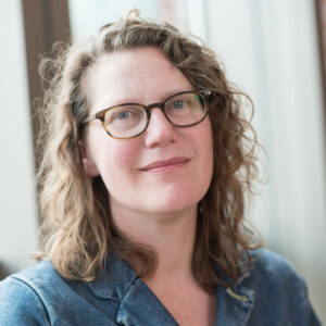 Portrait of Leontina Hormel. She has flowy brown hair, dark glasses and a teal shirt. 