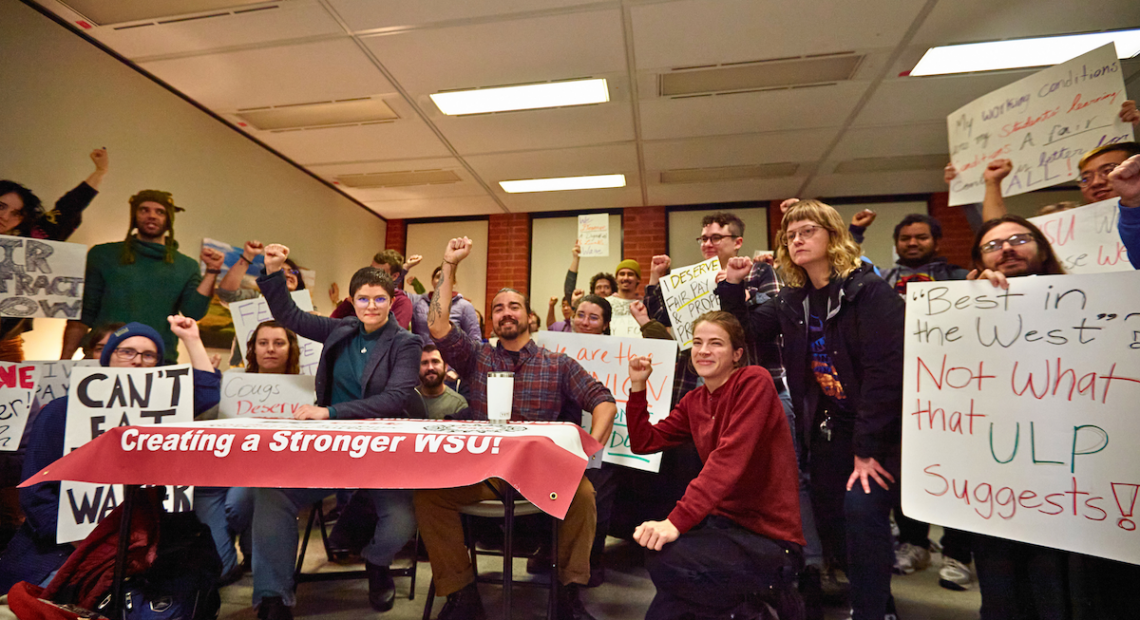 Students with handmade signs hold a rally around a table inside a building on the WSU campus. Many students are raising their fist in the air.