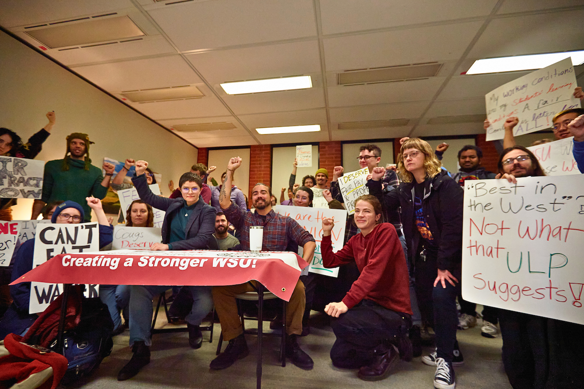 Students with handmade signs hold a rally around a table inside a building on the WSU campus. Many students are raising their fist in the air.