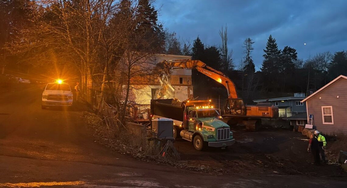 An orange excavator tears into a white three story house.