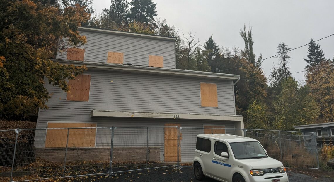 A three story dark white house with plywood in the windows sits surrounded by trees under a cloudy sky.