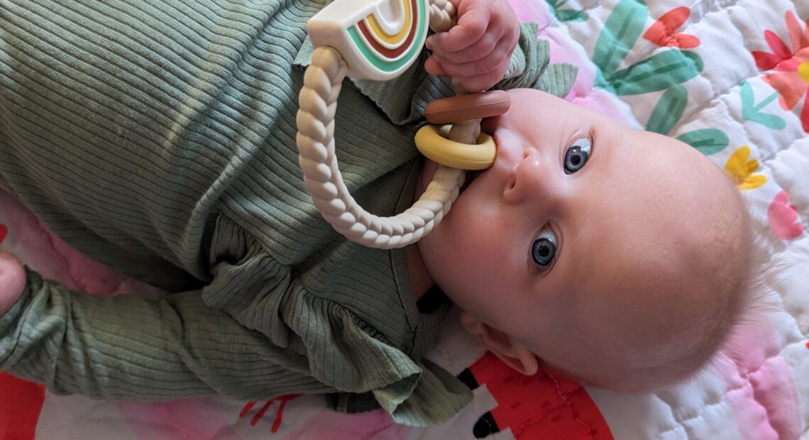 A baby in a green top holds a beige rattle toy to her month while laying on a pink blanket.