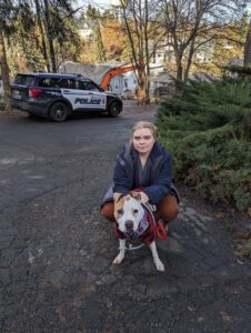 A girl with blonde hair sits with her white pitbull on the pavement.