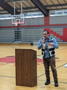 A Nez Perce man in braids and a vest stands before a wooden podium in a gymnasium.