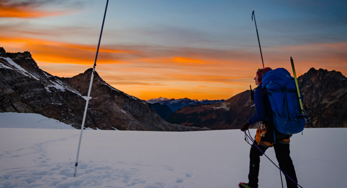 A woman in black snow pants and a yellow jacket with a big blue backpack measures snow with a large rod on top of a mountain covered in snow. An orange sunset is visible in the distance.