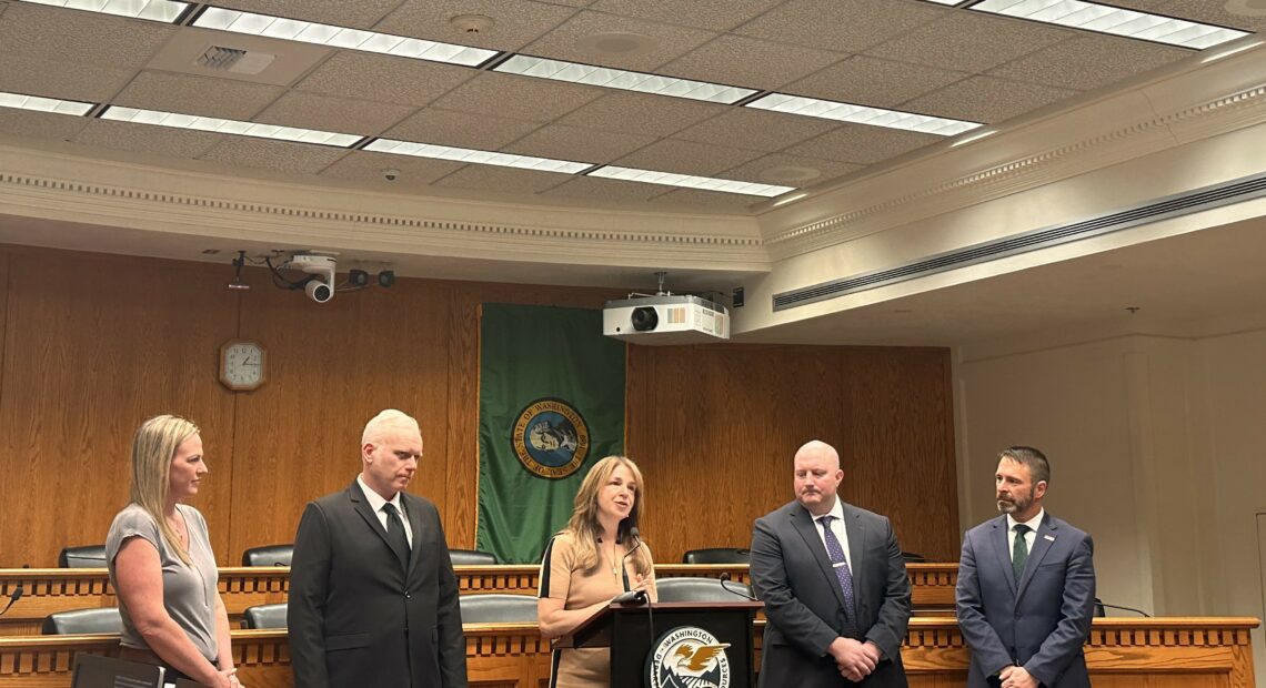 Five people in suits and dresses stand inside a conference room at the Capitol building in Olympia, Washington under fluorescent lights.