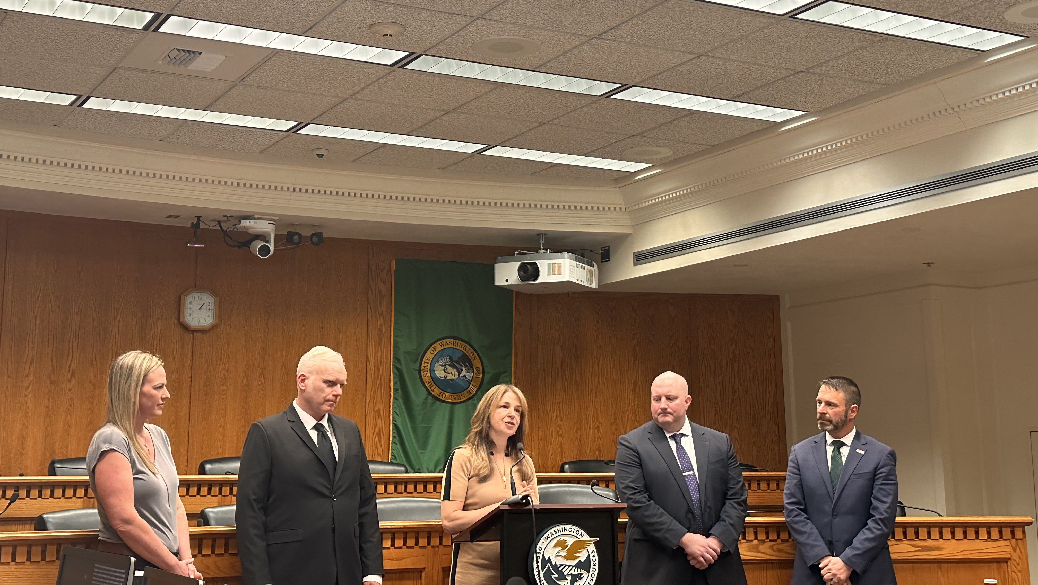 Five people in suits and dresses stand inside a conference room at the Capitol building in Olympia, Washington under fluorescent lights.