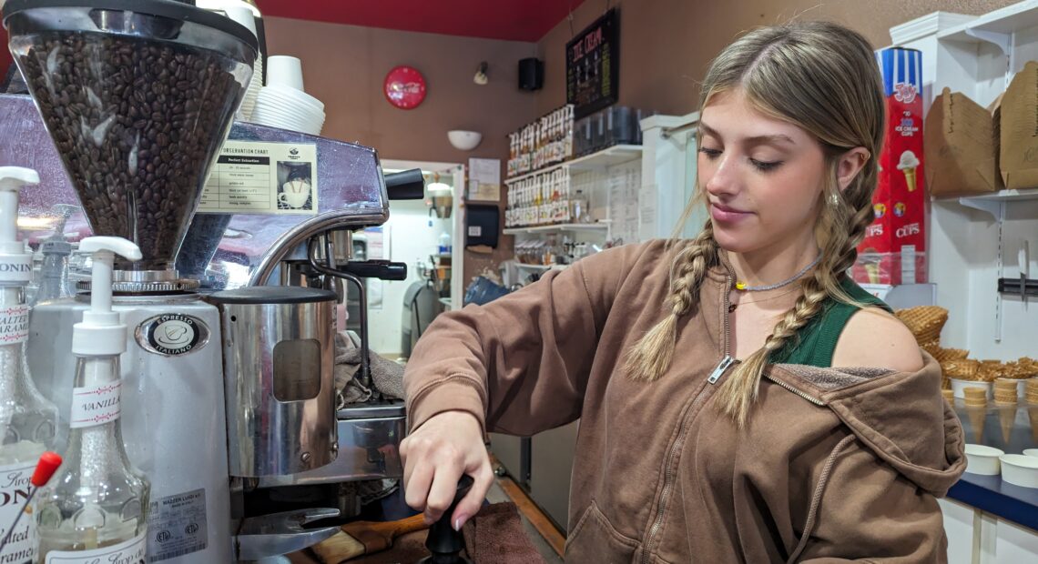 A woman with blonde hair and a brown sweatshirt pulls shots for an Americano at a silver espresso machine.