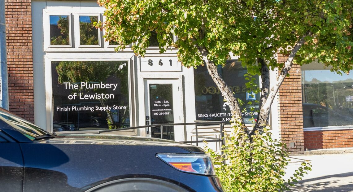 A black car is parked in the foreground. A building, the Plumbery of Lewiston, is pictured behind the car.