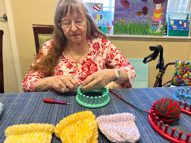 A woman with long gray hair and a white blouse with red flowers on it sits at a table with a blue tablecloth. She has a turquoise loom in her hands with red and green yarn tied to it. To the side, there is a red loom and a blue loom. In front of her are three completed hats. Two are yellow and one is purple.