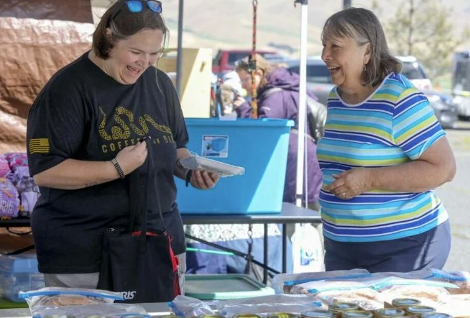 Two women stand in front of a table outside. The woman on the left is wearing a black shirt and is holding a wrapped package. The woman on the right wears a blue striped shirt.