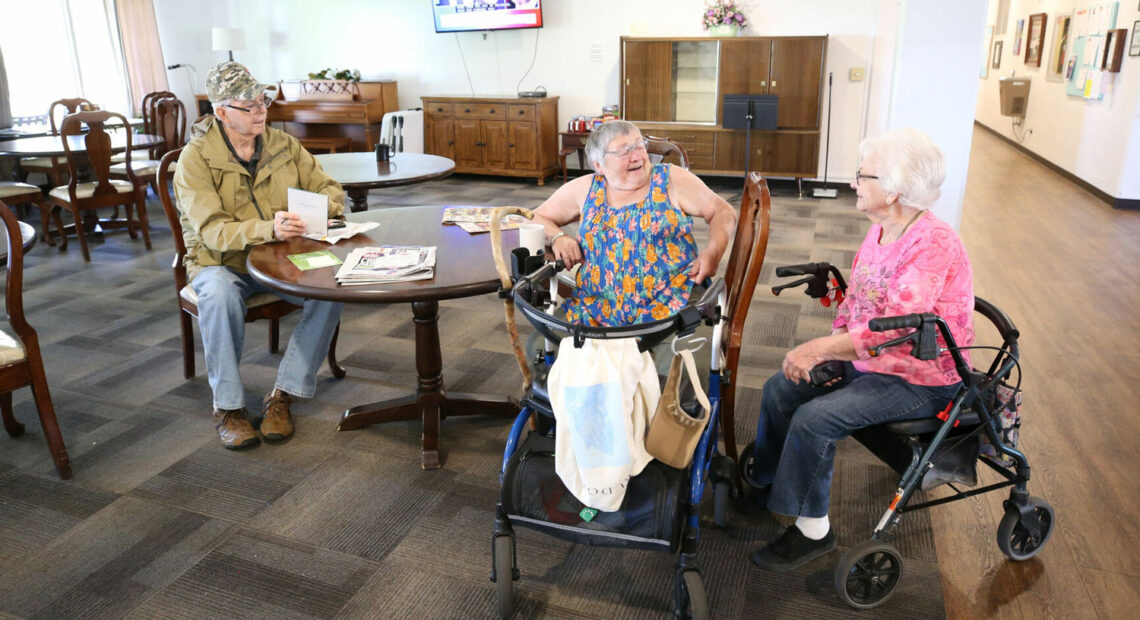 Three people are gathered around a circular, brown table having a conversation. A man in brown jacket is seated to the far left. A woman in a blue and pink checked shirt is seated to the right. A woman is seated in a walker next to her.