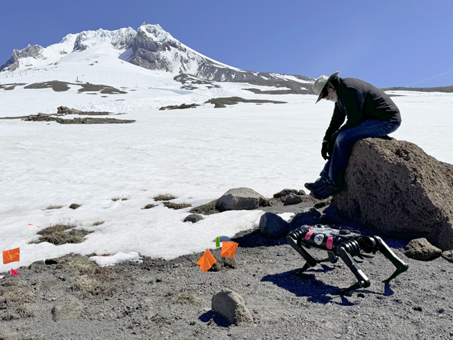 A man dressed in black sits on a gray rock. There is snow behind him. He is looking at a black robot in the shape of a dog. Behind the man and the robot is the summit of Mount Hood.