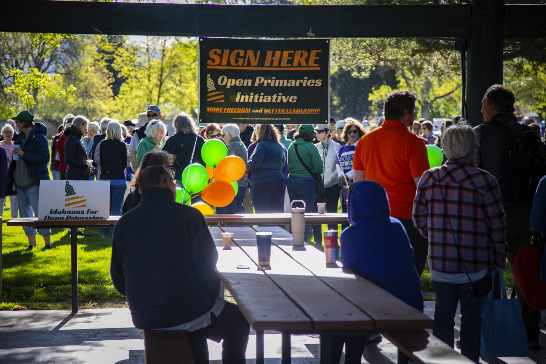 People stand in green grass and sit at a picnic table under a sign that reads "sign here."