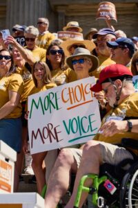 People in yellow t-shirts gather at the steps of the Boise State Capitol Building holding a sign that says "More Choice More Voice."