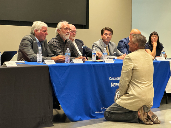 A man in a tan coat and tan pants is on his knees in front of a table with a blue table cloth. Six people, five men and one woman, are sitting at the table, staring at the man on his knees.