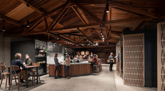 A bar with several people standing by it is pictured. The ceiling in the building is made of dark, exposed wood beams.