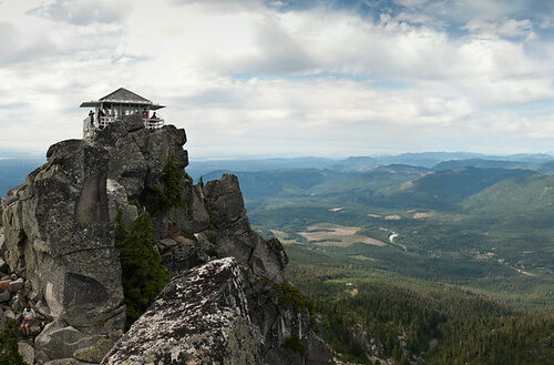 A small house with glass windows surrounding the entire outside sits on top of spiky gray rocks. In the distance are green hills all the way to the horizon. There are gray-white clouds in the sky.