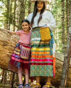 A woman and a girl in colorful outfits pose in front of a large log in the forest.