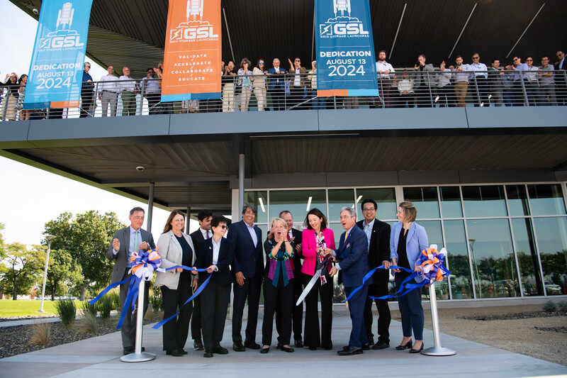 A group of people in suits stand in front of a building, where people are on a balcony. One woman in pink is holding giant scissors to cut a ribbon.