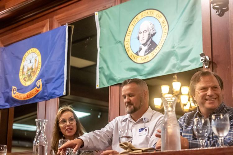 A woman and two men sit at a table with water glasses in front of them. The state flags of Idaho and Washington are behind them.