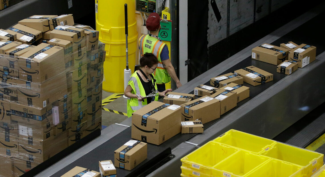 Packages move down a conveyor belt. Two people in high-visibility vests stand next to the conveyor belt.