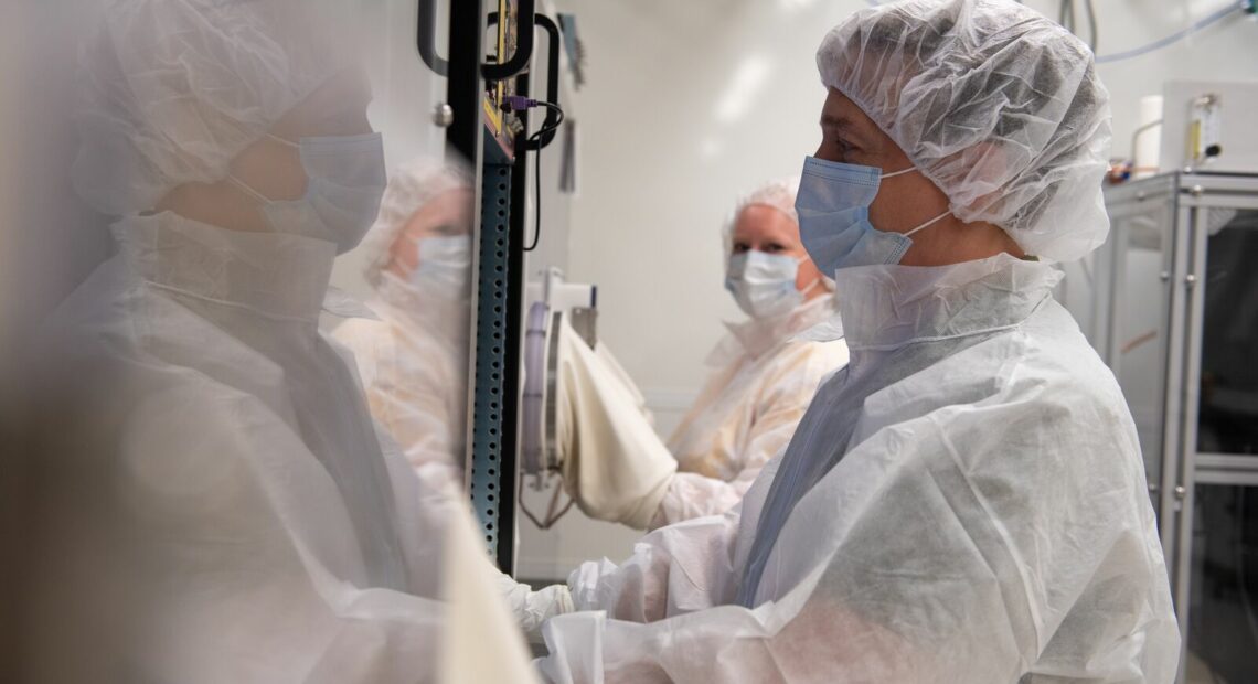 A man in a white lab coat, a white hair net and a blue mask is surrounded by other people, who are blurry in the photo, but also appear to be wearing lab coats and masks. He is standing in front of a glass covered box.