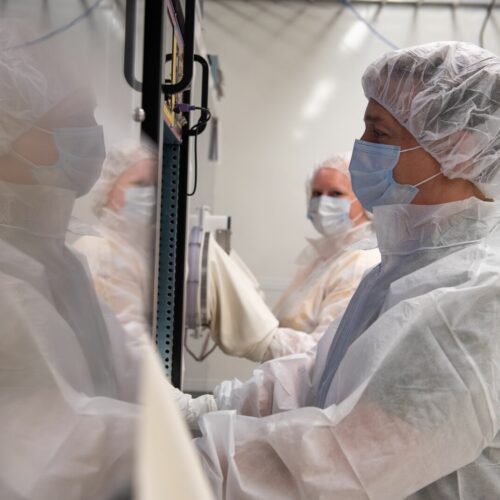 A man in a white lab coat, a white hair net and a blue mask is surrounded by other people, who are blurry in the photo, but also appear to be wearing lab coats and masks. He is standing in front of a glass covered box.