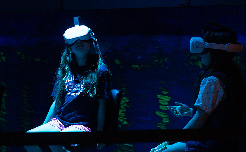 Two girls with virtual reality headsets are in a dark room. One of them is looking up towards the ceiling.