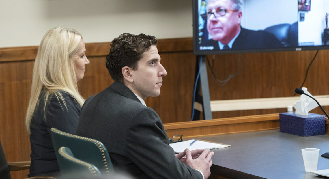 A woman and a man sit at a black table in a courtroom. A TV screen, with the judge pictured, is to the left of them.