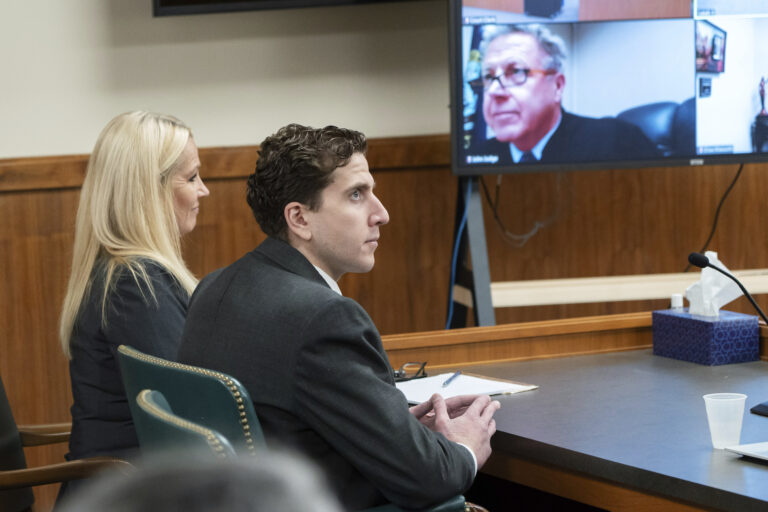 A woman and a man sit at a black table in a courtroom. A TV screen, with the judge pictured, is to the left of them.