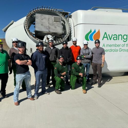 Ten men wearing hard hats stand in front of the top part of a turbine that is lying on its side on the ground. There is a sign on the equipment that says "Avangrid." They are standing on cement. The sky is blue in the background.