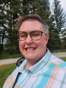 A portrait of Julia Keleher. They are wearing a blue and orange plaid shirt and black glasses. Evergreen trees are pictured in the background.