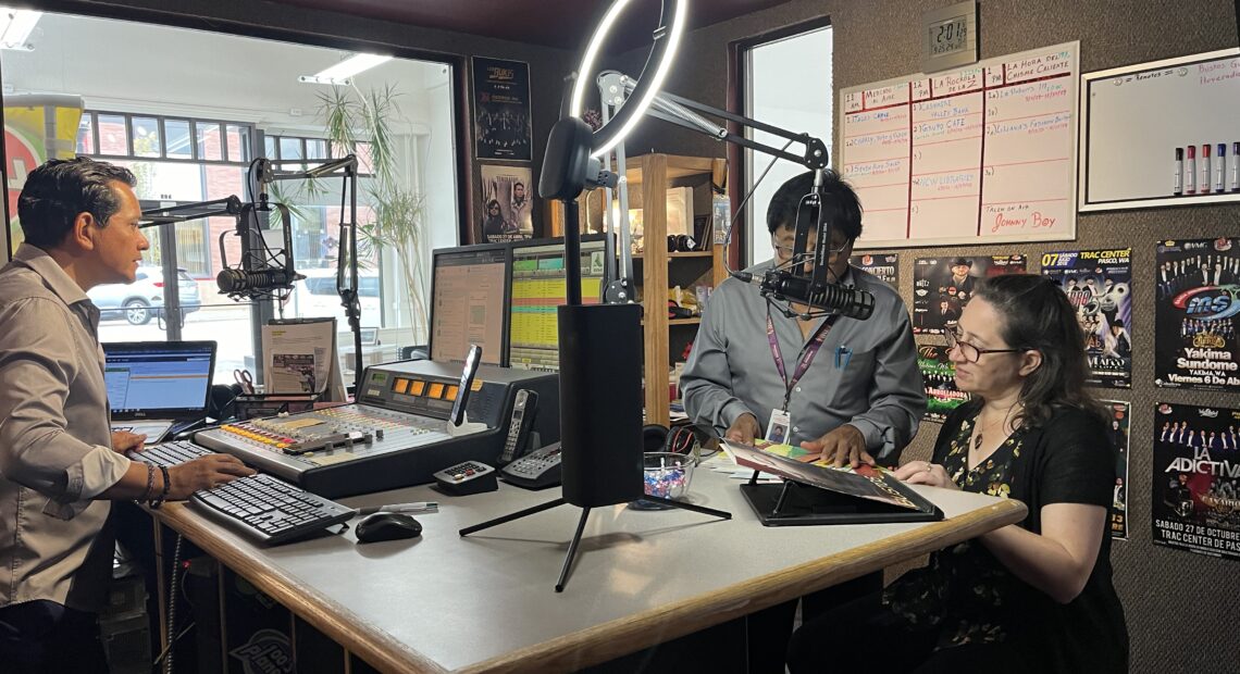 Two men and a woman gather in a sound booth in a radio station.