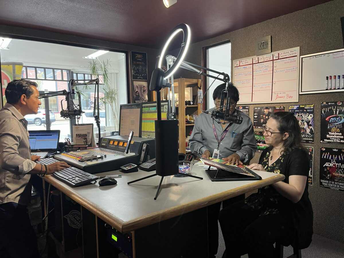 Two men and a woman gather in a sound booth in a radio station.