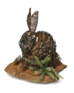 A bunny made out of stick-looking wire. It has spine needles at its feet and is on top of a brown stone.