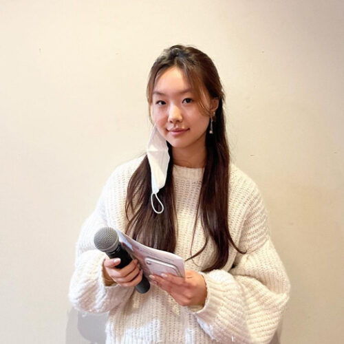 A young woman with long black hair is holding a microphone and a cellphone. She is wearing a white sweater and standing against a white background.