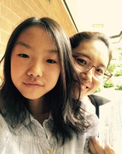 A young girl in a shite button up shirt is in front of her mom, who is wearing glasses. There is a brick wall behind them.