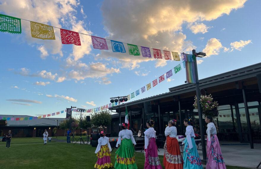 Six dancers, in brightly colored skirts, stand in a grassy area. A bright blue sky with clouds is above them.