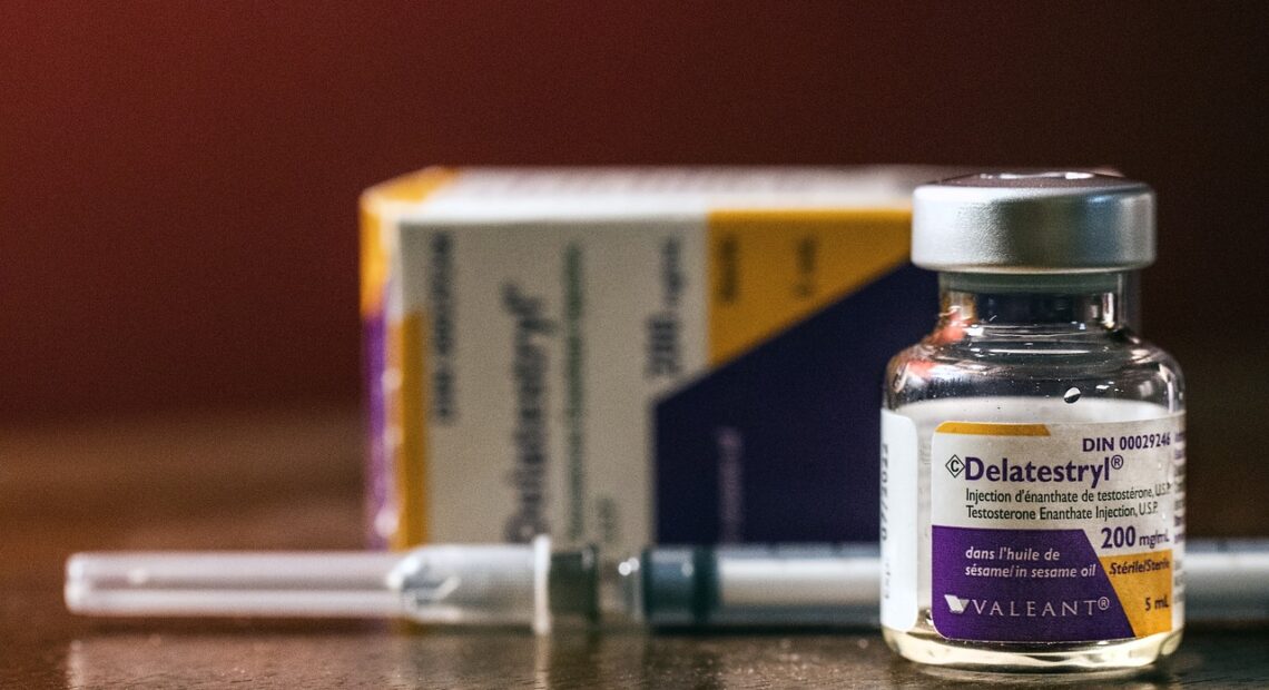 A vial of testosterone enanthate, with a needle behind it, sits on a table.