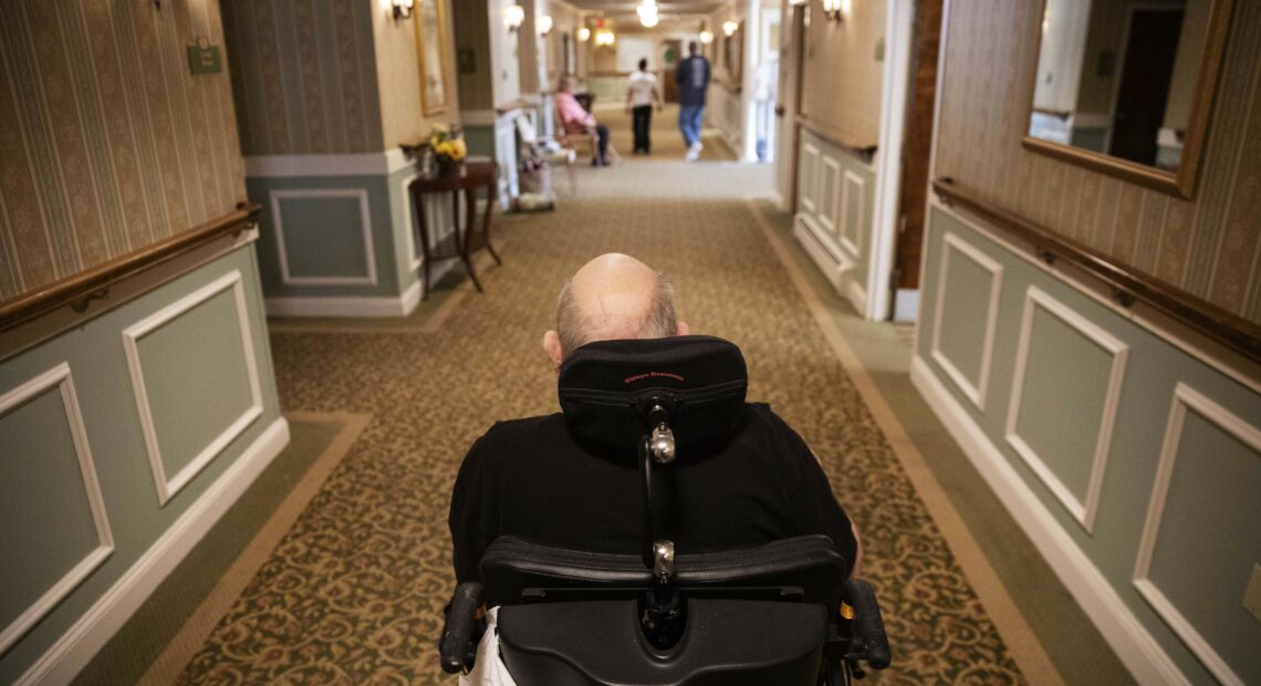 Elderly man in wheelchair in a nursing home hallway.