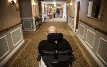 Elderly man in wheelchair in a nursing home hallway.