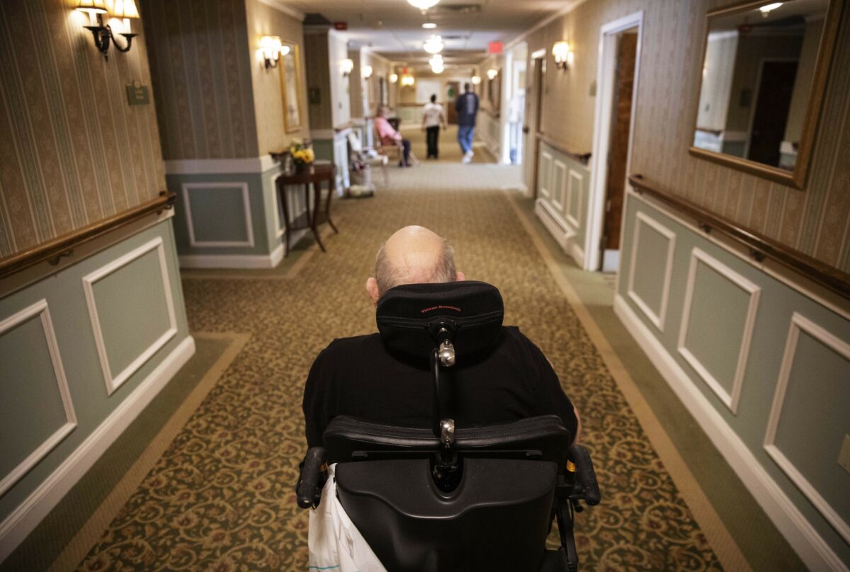 Elderly man in wheelchair in a nursing home hallway.