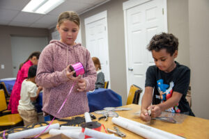 A girl in a pink sweatshirt is holding a pink ribbon. She is standing behind a table that's covered in construction paper. A boy in a black shirt is sitting at the table. They are standing in front of several white doors.