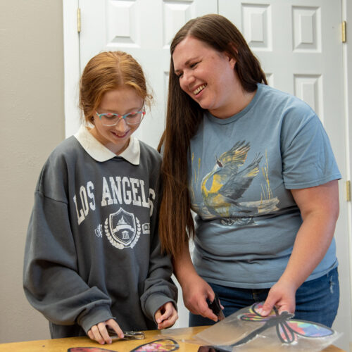 A young girl in a gray sweatshirt with a white collar stands next to a woman in a blue shirt. The girl has red hair. The woman has brown hair. They are standing behind a plastic table and in front of a white door. The woman is smiling at the girl.