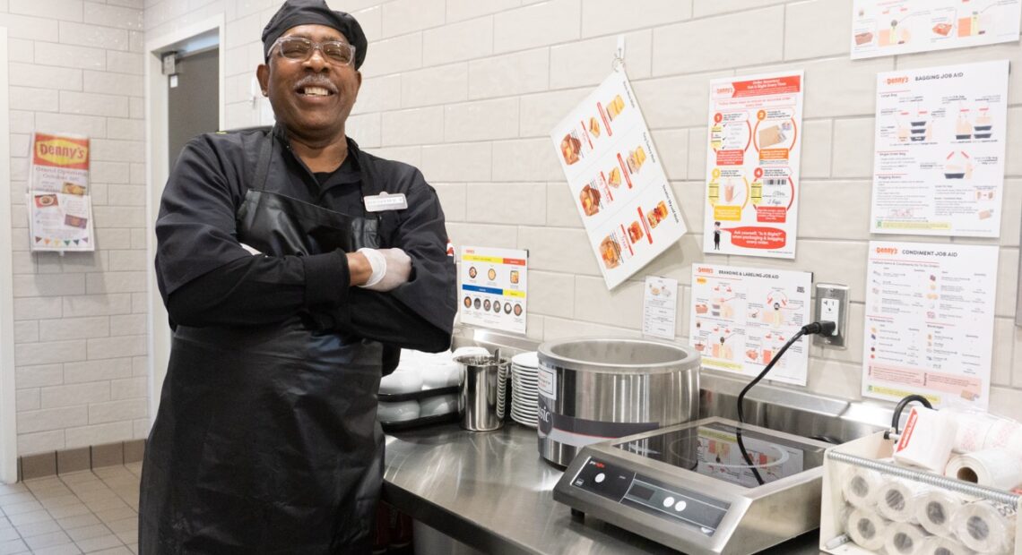 A man in a black chef's outfit stands in a kitchen.