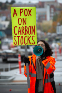 A student wearing a high-visibility jacket speaks into a blue megaphone. She is holding a yellow sign that reads "A pox on carbon stocks."