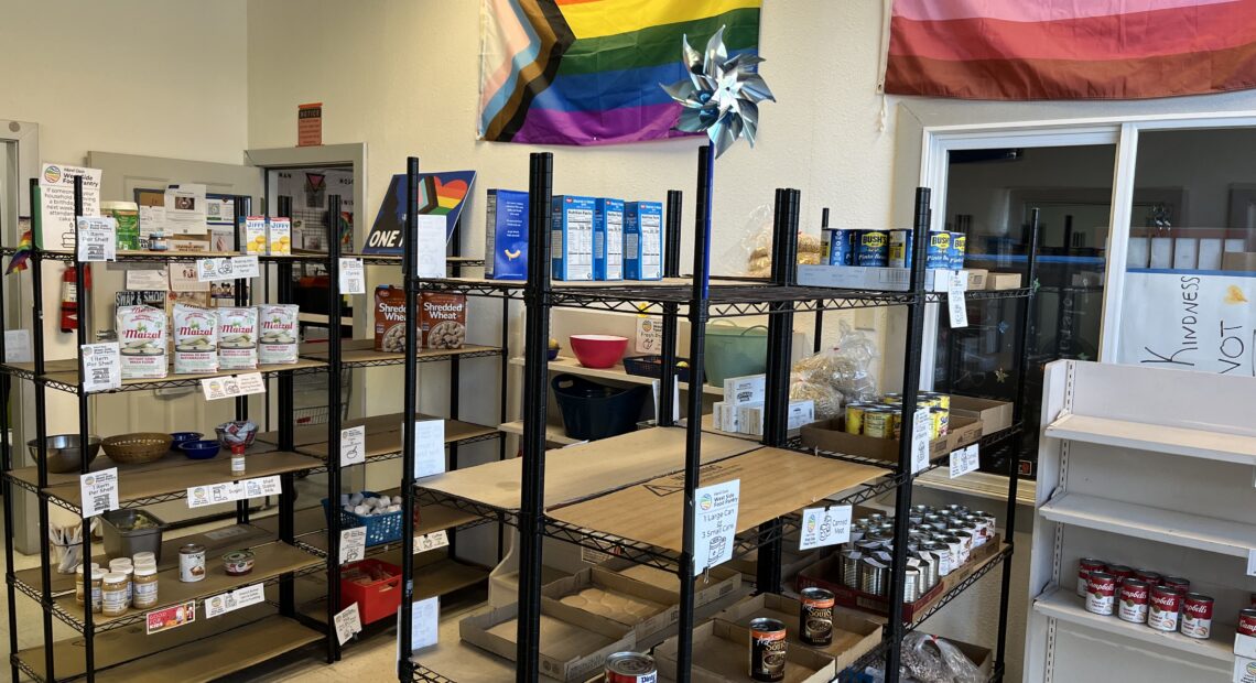 Several racks of canned goods are pictured in a room. Above the racks is a pride flag.